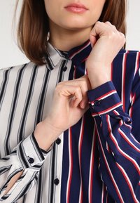 Niebieska i biała koszula z kołnierzykiem w pionowe czerwone paski. Posiada guziki na rękawach i mankietach, pokazując połączenie materiałów.