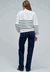Sudadera blanca con rayas horizontales en las mangas de color azul marino, combinada con jeans de color azul oscuro. La sudadera tiene un cuello redondo y un corte relajado.