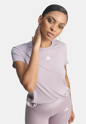 Donna che indossa un completo sportivo Adidas viola chiaro con il logo, posando con una mano che le sfiora il collo su uno sfondo bianco.