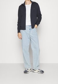 Uomo che indossa una giacca zip blu navy, una maglietta bianca, jeans larghi di un blu chiaro e sneakers bianche e blu navy, in piedi con una mano in tasca.
