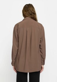 Chemise longue à manches longues de couleur marron avec une coupe décontractée, une légère brillance, un pli dans le dos et des poignets boutonnés. Le matériau semble léger et lisse.