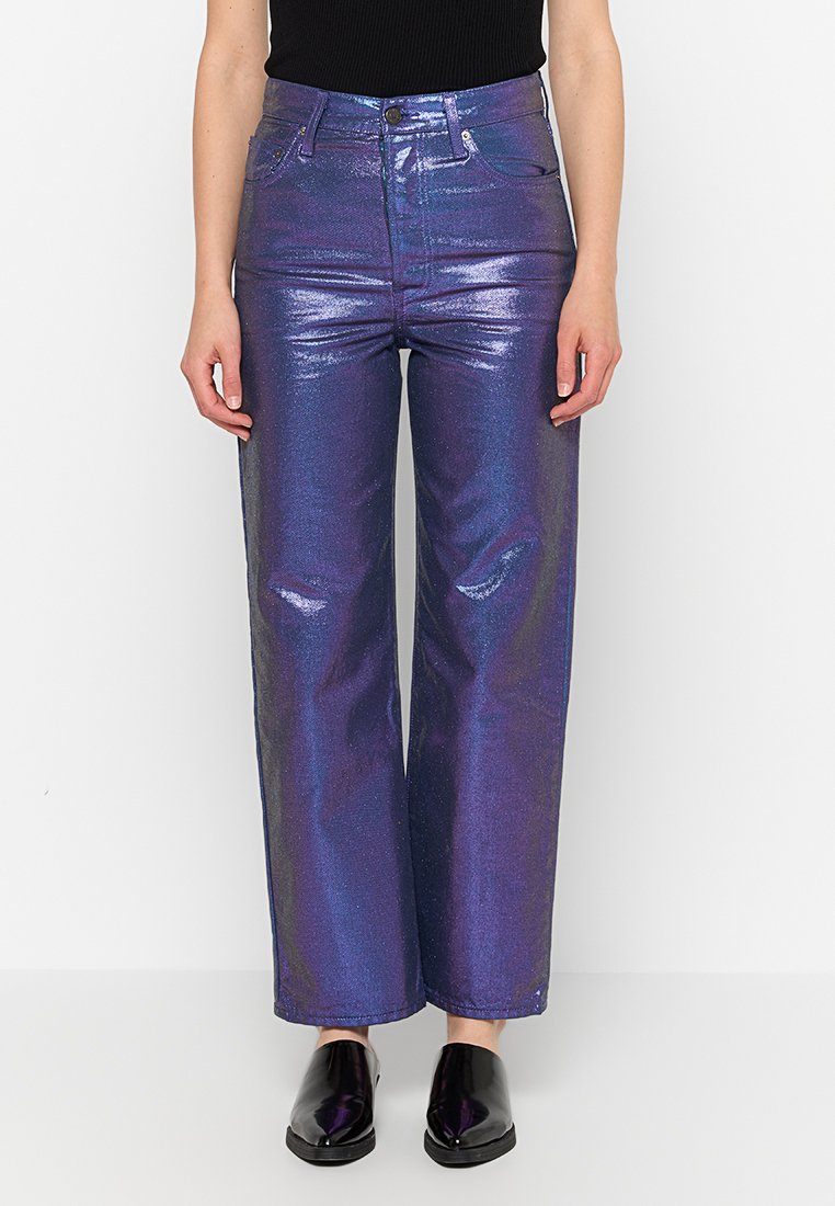 Persona con pantalones rectos brillantes de cintura alta en color púrpura, top sin mangas negro y zapatos negros tipo slip-on de pie contra un fondo blanco.
