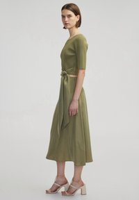 Ensemble deux pièces en vert olive comprenant un haut à manches courtes côtelé et une jupe fluide, nouée à la taille avec une ceinture en tissu. Talons épais.