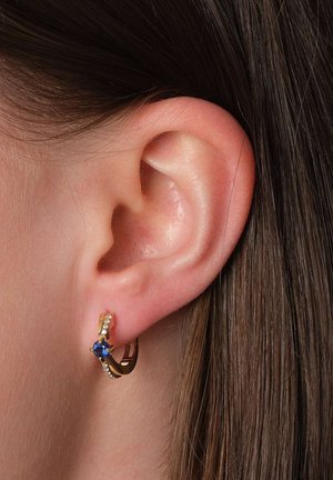 Ear with gold hoop earring featuring a blue gemstone and small clear stones, set against brown hair and skin background.