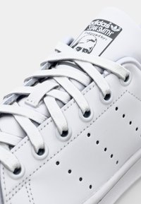 Biały skórzany sneaker z płaskimi białymi sznurowadłami, perforowanymi detalami z boku oraz oznaczoną metką na języku z ilustracją twarzy.