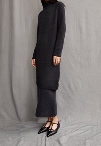 Robe tricotée gris foncé à manches longues et col montant, avec une fente latérale et une texture côtelée. Associée à des ballerines pointues noires.