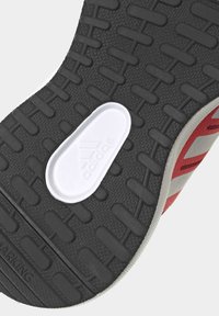Černá gumová podrážka s texturovaným vzorem, obsahující bílý polstrovaný prvek s logem Adidas a červené designové akcenty na boku.