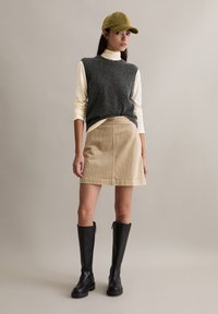 Grijze wollen mouwloze trui, beige mini-rok van ribfluweel en zwarte kniehoge laarzen. Olijfgroene pet voegt accent toe. Zachte texturen, neutraal palet.