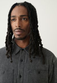 Camisa de botones en blanco y negro con cuello, dos bolsillos en el pecho y mangas cortas, con tela texturizada y botones contrastantes.