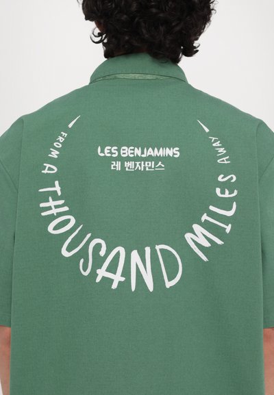 Zelena majica kratkih rukava s bijelim natpisom "LES BENJAMINS" i kružnim frazama "FROM A THOUSAND MILES AWAY" u različitim fontovima.