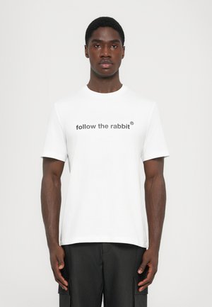 T-shirt en coton blanc avec un col rond et des manches courtes. Présente le texte "suivez le lapin®" en une police grise atténuée sur la poitrine.