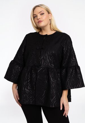 Frau in einer strukturierten, schwarzen Bluse mit langen Ärmeln und auslaufenden Manschetten sowie schwarzen Hosen, die vor einem weißen Hintergrund steht.