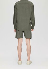 Man in olijfgroene lange mouwen shirt en bijpassende shorts, staande met zijn rug naar de camera op een effen lichte achtergrond.