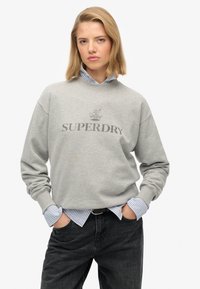 Szary sweatshirt z napisem "SUPERDRY", z prążkowanym dekoltem i opuszczonymi ramionami, założony na niebieską i białą koszulę w paski, zestawiony z czarnymi dżinsami.