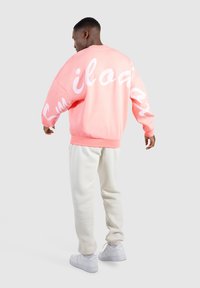 Sudadera rosa de gran tamaño con texto blanco en las mangas, combinada con pantalones de chándal beige claro y zapatillas blancas, vista desde atrás.