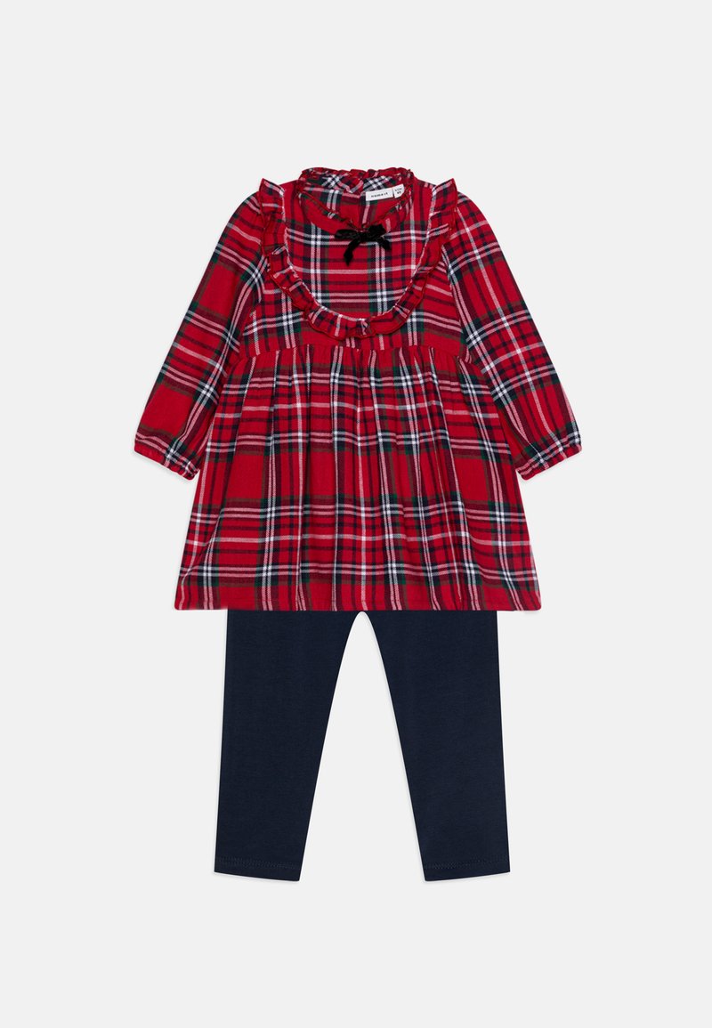 Piros kockás ruha hosszú ujjakkal, fodros nyakkivágással és fekete masnival. Sötétkék leggingssel párosítva, mindkettő puha anyagból készült.