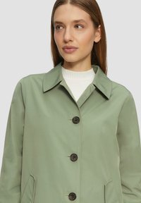 Lichtgroene button-up jas met een kraag, vier knopen aan de voorkant en zijzakken. De stof heeft een soepele textuur en is licht van gewicht.