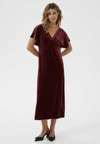 Kvinna iklädd en vinröd sammet V-ringad midiklänning med korta ärmar och svarta spetsiga klackar, stående med händerna bakom ryggen.