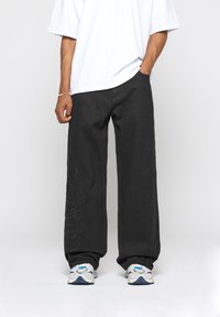 Pantalon en denim noir à jambes larges avec un motif de feuilles en relief sur un côté, associé à un t-shirt blanc ample et des baskets gris et bleu.