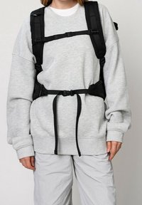 Personne portant un sweat-shirt gris clair et un pantalon avec une sangle noire de type harnais fixée sur la poitrine et la taille.