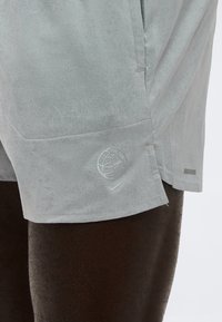 Shorts de sport gris clair portés par une personne, avec un globe et les logos Nike Swoosh près de l'ourlet sur la jambe gauche.