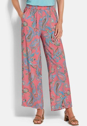 Vrouw draagt roze broek met wijde pijpen met blauwgroene paisley patronen, gecombineerd met beige sandaal met hak en een lichtblauwe top.