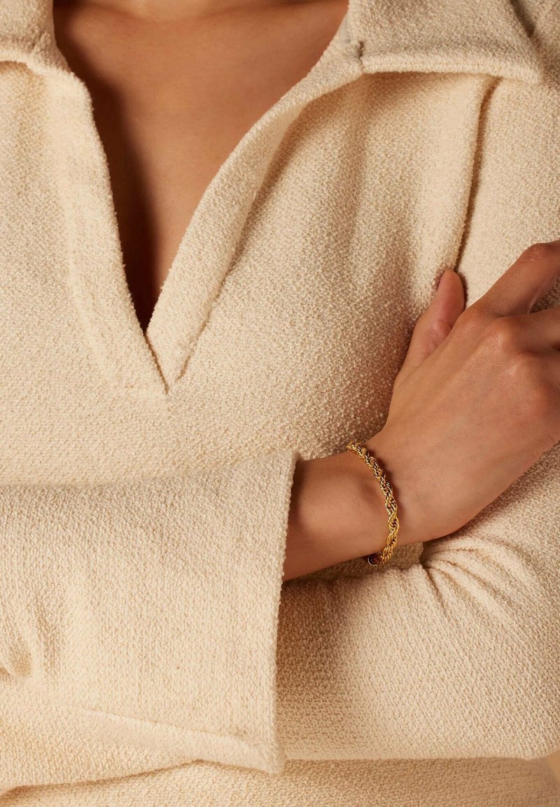 Gouden gedraaide kettingarmband om een pols, gecombineerd met een gestructureerde beige top met een V-hals en brede mouwen.