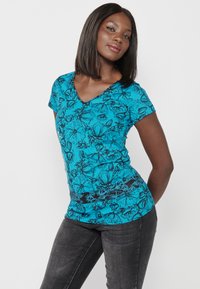 T-shirt turquoise à col V avec imprimé floral noir, manches courtes et design ajusté ; associé à un jean sombre.