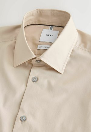 Beige bomulds knap-op skjorte med spids krave, mærket "NEXT" og "Cotton Street", pænt foldet mod en hvid baggrund.