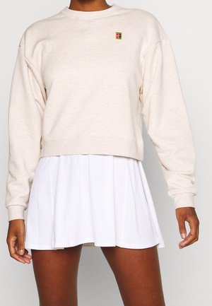 Person trägt einen beigefarbenen, langärmligen, kurzgeschnittenen Pullover mit einem kleinen, bunten Logo und einen weißen, plissierten Rock.