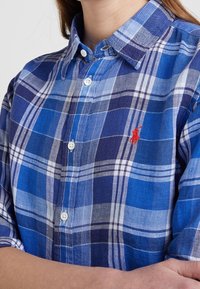 Blå rutig skjorta med knappar, tillverkad av bomull, med vita och marinblå ränder, en klassisk krage och en röd logotyp på bröstet.