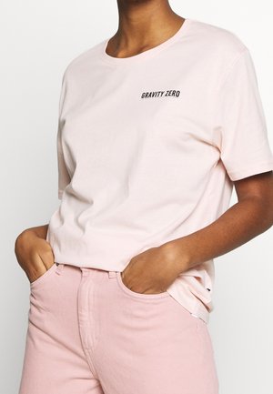 Person trägt ein hellrosa kurzärmeliges T-Shirt mit dem Schriftzug „GRAVITY ZERO“ auf der Brust und passende rosa Hosen, die Hände in den Taschen.