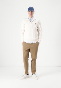 Witte kabelgebreide trui met een rits, lichtblauw overhemd met kraag eronder, beige broek en witte sneakers. Blauwe pet als accessoire.