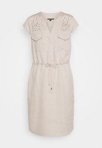 Robe beige avec un décolleté en V, des manches courtes, deux poches poitrine et des clous décoratifs sur les épaules. Taille froncée avec un détail de cordon.