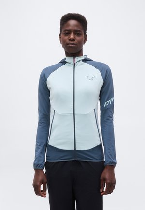 Személy, aki világoskék és sötétkék cipzáras sportdzsekit kapucnival visel, egyszerű fehér háttér előtt áll.