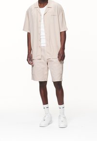 Ljusbeige kortärmad skjorta med knappar och präglad text, parat med matchande cargoshorts. Vita strumpor och sneakers fulländar looken.