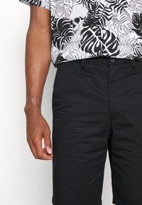 Black shorts with a smooth texture, paired with a white short-sleeve shirt featuring a bold black leaf and skull pattern.