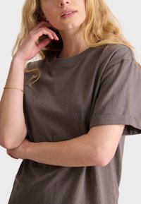 Brown cotton t-shirt with a round neckline, short sleeves, and a smooth texture, worn by a person with wavy hair.