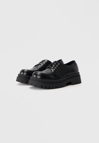 Pantofi din piele neagră, cu șireturi, cu talpă groasă și zimțată, cu vârfuri rotunde și gauri pentru șireturi din metal, prezentati pe un fundal alb simplu.