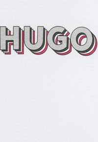 Hvit T-skjorte med ordet "HUGO" i 3D-effekt bokstaver. Bokstavene er sølv med rosa og svarte aksenter, som reflekterer en teksturert overflate.