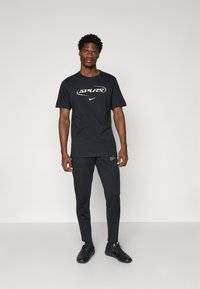 Sort atletisk T-shirt med "SPURS" logo og Nike swoosh. Kombineret med sorte tætsiddende bukser og sorte sneakers. Glat stof tekstur.