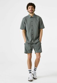 Camisa de manga corta con cuello y pantalones cortos a juego en una tela texturizada de color verde oscuro. Presenta cierre de botones en la parte delantera, bolsillos laterales y un corte relajado.