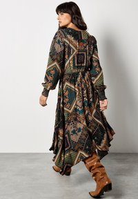 Lange, vloeiende jurk in een patchworkpatroon met aardetinten, voorzien van lange mouwen, een gegatherde taille en een ontspannen pasvorm, gecombineerd met kniehoog laarzen.