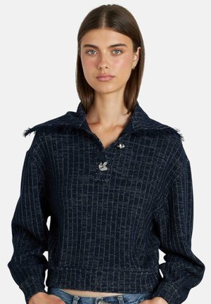 Junge Frau mit glattem braunem Haar, die einen dunkelblauen, gemusterten Strickpullover mit übergroßem Kragen und silbernen Knöpfen trägt.