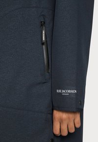 Chaqueta azul marino con una textura suave, que cuenta con un bolsillo con cremallera, un logo en la manga y detalles de costura en contraste.