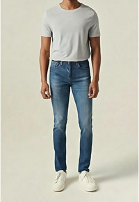 Homme portant un t-shirt à manches courtes gris clair, un jean slim bleu et des baskets blanches décontractées, debout devant un fond uni.