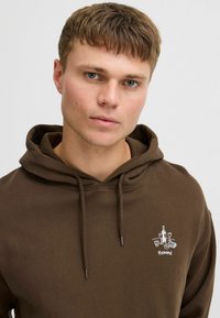 Brauner Hoodie mit Kordelzugkapuze, ausgestattet mit einer kleinen gestickten Grafik von Flaschen und Gläsern, aus weichem Material mit glatter Textur.