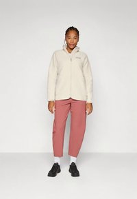 Femeie în picioare, purtând o jachetă de fleece crem cu fermoar, pantaloni de culoare roz, șosete albe și adidași negri, pozând într-un fundal alb simplu.