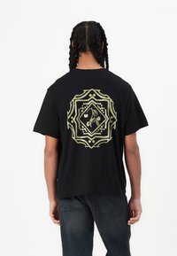 Sort bomulds t-shirt med et stort guld geometrisk design med et kirsebærmotiv på bagsiden. Korte ærmer og en afslappet pasform.