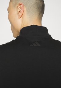 Juodas polo marškinėliai su aukštu apykakle, pagaminti iš lygaus audinio. Ant nugaros yra diskretiškas juodas Adidas logotipas.